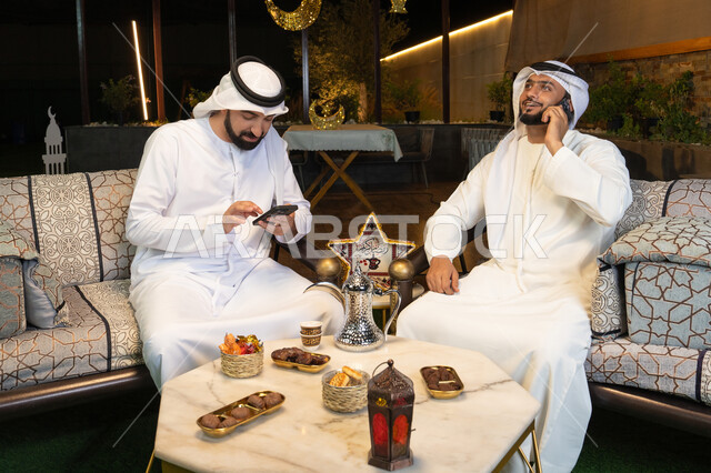 اجراء مكالمة هاتفية باستخدام الهاتف المحمول بتعابير السرور، زينة واضاءة رمضانية، الجلسة الليلة الخارجية في احدى شاليهات الامارات، حسن الاستقبال وكرم الضيافة في شهر رمضان المبارك، صديقان عربيان خليجيان اماراتيان يرتديان الكندورة والغترة يجلسان في الهواء الطلق