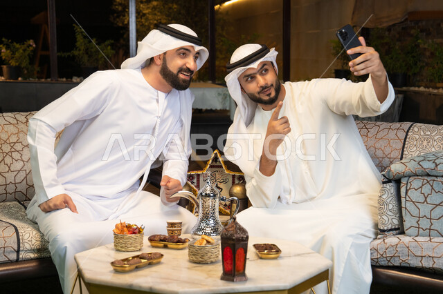 الاستقبال وكرم الضيافة في شهر رمضان المبارك، توثيق اللحظات السعيدة،  صديقان عربيان خليجيان اماراتيان يرتديان الكندورة والغترة يجلسان في الهواء الطلق، التقاط صور سيلفي باستخدام الهاتف المحمول بتعابير السرور، زينة واضاءة رمضانية، الجلسة الليلة الخارجية