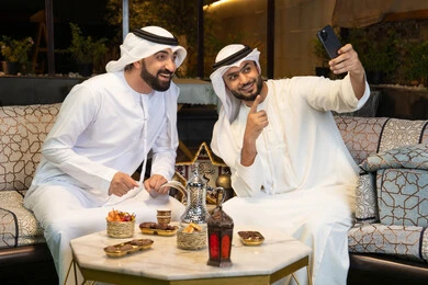 Warm welcome and hospitality during the holy month of Ramadan, documenting happy moments, two Emirati Gulf Arab friends wearing kandura and ghutra sitting outdoors, taking selfies using mobile phones with happy expressions, Ramadan decorations and lighting, outdoor night session