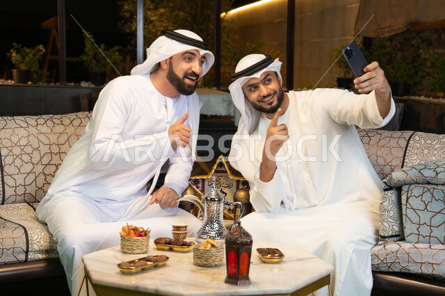توثيق اللحظات السعيدة، حسن الاستقبال وكرم الضيافة في شهر رمضان المبارك، صديقان عربيان خليجيان اماراتيان يرتديان الكندورة والغترة يجلسان في الهواء الطلق، التقاط صور سيلفي باستخدام الهاتف المحمول بتعابير السرور، زينة واضاءة رمضانية، الجلسة الليلة الخارجية