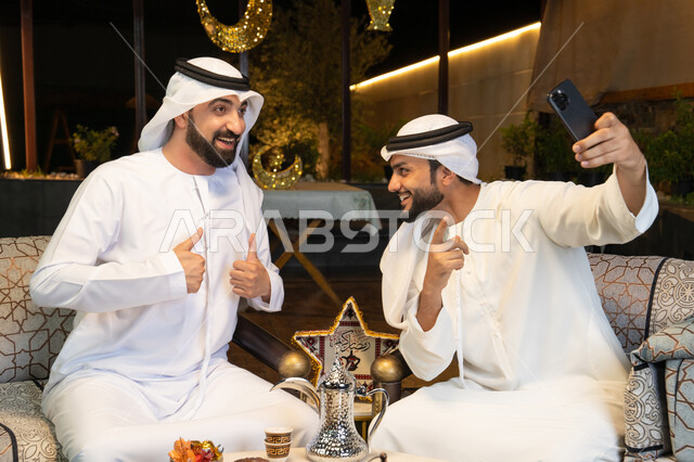 حسن الاستقبال وكرم الضيافة في شهر رمضان المبارك، توثيق اللحظات السعيدة،  صديقان عربيان خليجيان اماراتيان يرتديان الكندورة والغترة يجلسان في الهواء الطلق، التقاط صور سيلفي باستخدام الهاتف المحمول بتعابير السرور، زينة واضاءة رمضانية، الجلسة الليلة الخارجية