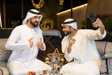 حسن الاستقبال وكرم الضيافة في شهر رمضان المبارك، توثيق اللحظات السعيدة،  صديقان عربيان خليجيان اماراتيان يرتديان الكندورة والغترة يجلسان في الهواء الطلق، التقاط صور سيلفي باستخدام الهاتف المحمول بتعابير السرور، زينة واضاءة رمضانية، الجلسة الليلة الخارجية