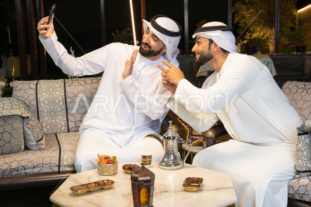 توثيق اللحظات السعيدة، حسن الاستقبال وكرم الضيافة في شهر رمضان المبارك، صديقان عربيان خليجيان اماراتيان يرتديان الكندورة والغترة يجلسان في الهواء الطلق، التقاط صور سيلفي باستخدام الهاتف المحمول بتعابير السرور، زينة واضاءة رمضانية، الجلسة الليلة الخارجية
