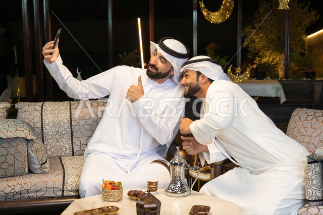 حسن الاستقبال وكرم الضيافة في شهر رمضان المبارك، توثيق اللحظات السعيدة،  صديقان عربيان خليجيان اماراتيان يرتديان الكندورة والغترة يجلسان في الهواء الطلق، التقاط صور سيلفي باستخدام الهاتف المحمول بتعابير السرور، زينة واضاءة رمضانية، الجلسة الليلة الخارجية