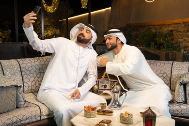 توثيق اللحظات السعيدة، حسن الاستقبال وكرم الضيافة في شهر رمضان المبارك، صديقان عربيان خليجيان اماراتيان يرتديان الكندورة والغترة يجلسان في الهواء الطلق، التقاط صور سيلفي  باستخدام الهاتف المحمول بتعابير السرور، زينة واضاءة رمضانية، الجلسة الليلة الخارجية