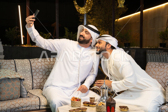 Documenting happy moments, warm welcome and hospitality during the holy month of Ramadan, two Emirati Gulf Arab friends wearing kandura and ghutra sitting outdoors, taking selfies using mobile phones with happy expressions, Ramadan decorations and lighting, outdoor night session