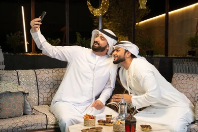 Documenting happy moments, warm welcome and hospitality during the holy month of Ramadan, two Emirati Gulf Arab friends wearing kandura and ghutra sitting outdoors, taking selfies using mobile phones with happy expressions, Ramadan decorations and lighting, outdoor night session