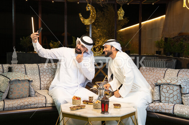 Documenting happy moments, warm welcome and hospitality during the holy month of Ramadan, two Emirati Gulf Arab friends wearing kandura and ghutra sitting outdoors, taking selfies using mobile phones with happy expressions, Ramadan decorations and lighting, outdoor night session