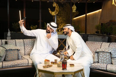 توثيق اللحظات السعيدة، حسن الاستقبال وكرم الضيافة في شهر رمضان المبارك، صديقان عربيان خليجيان اماراتيان يرتديان الكندورة والغترة يجلسان في الهواء الطلق، التقاط صور سيلفي  باستخدام الهاتف المحمول بتعابير السرور، زينة واضاءة رمضانية، الجلسة الليلة الخارجية