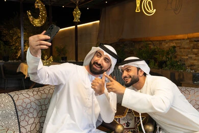 تصوير اللحظات السعيدة، حسن الاستقبال وكرم الضيافة في شهر رمضان المبارك، صديقان عربيان خليجيان اماراتيان يرتديان الكندورة والغترة يجلسان في الهواء الطلق، التقاط صور سيلفي  باستخدام الهاتف المحمول بتعابير السرور، زينة واضاءة رمضانية، الجلسة الليلة الخارجية