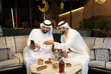 Gestures indicating joy and happiness, warm welcome and hospitality in the holy month of Ramadan, two Emirati Gulf Arab friends wearing kandura and ghutra sitting outdoors, Ramadan decorations and lighting, outdoor night session