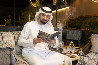 Reading, reciting and memorizing the Holy Quran, an Emirati Gulf Arab man wearing a kandura and ghutra reading the Holy Quran outdoors, reciting the Book of God Almighty, diligence and humility in worship, getting closer to the Lord of the Worlds in the blessed month of Ramadan, Ramadan decorations and lighting
