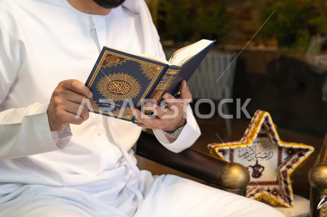 قراءة وتلاوة وحفظ القرآن الكريم، رجل عربي خليجي اماراتي يرتدي الكندورة والغترة  يقوم بقراءة المصحف الشريف في الهواء الطلق بالخارج، تلاوة كتاب الله تعالى ,الإجتهاد و الخشوع في العباده, التقرب الى رب العالمين في شهر رمضان المبارك، زينة واضاءة رمضانية