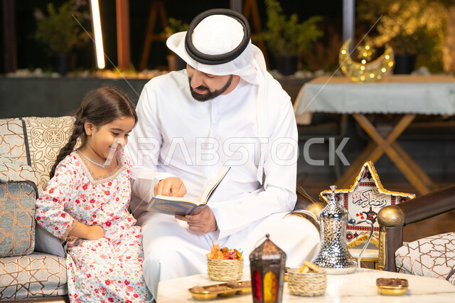 Reading, reciting and memorizing the Holy Quran, an Emirati Gulf Arab man wearing a kandura and ghutra holding the Holy Quran sitting with his daughter in the open air outside, reciting and reading the Book of God Almighty, diligence and humility in worship, getting closer to the Lord of the Worlds in the blessed month of Ramadan, Ramadan decorations and ighting