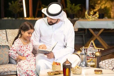 شرح وتلاوة وحفظ القرآن الكريم، رجل عربي خليجي اماراتي يرتدي الكندورة والغترة  يمسك المصحف الشريف يجلس مع طفلته في الهواء الطلق في الخارج، تلاوة وقراءة كتاب الله تعالى ,الإجتهاد و الخشوع في العباده, التقرب الى رب العالمين في شهر رمضان المبارك، زينة واضاءة رمضانية