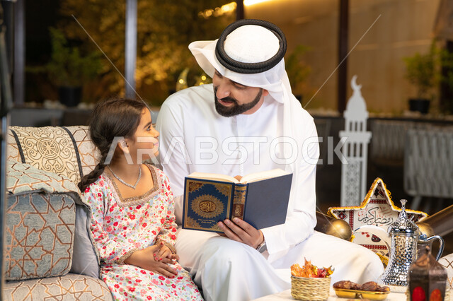قراءة وتلاوة وحفظ القرآن الكريم، رجل عربي خليجي اماراتي يرتدي الكندورة والغترة  يمسك المصحف الشريف يجلس مع طفلته في الهواء الطلق في الخارج، تلاوة وقراءة كتاب الله تعالى ,الإجتهاد و الخشوع في العباده, التقرب الى رب العالمين في شهر رمضان المبارك، زينة واضاءة رمضانية