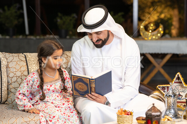 شرح وتلاوة وحفظ القرآن الكريم، رجل عربي خليجي اماراتي يرتدي الكندورة والغترة  يمسك المصحف الشريف يجلس مع طفلته في الهواء الطلق في الخارج، تلاوة وقراءة كتاب الله تعالى ,الإجتهاد و الخشوع في العباده, التقرب الى رب العالمين في شهر رمضان المبارك، زينة واضاءة رمضانية