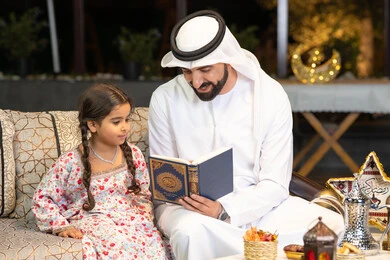 شرح وتلاوة وحفظ القرآن الكريم، رجل عربي خليجي اماراتي يرتدي الكندورة والغترة  يمسك المصحف الشريف يجلس مع طفلته في الهواء الطلق في الخارج، تلاوة وقراءة كتاب الله تعالى ,الإجتهاد و الخشوع في العباده, التقرب الى رب العالمين في شهر رمضان المبارك، زينة واضاءة رمضانية