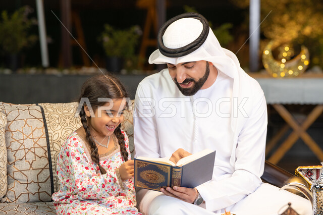 Reading, reciting and memorizing the Holy Quran, an Emirati Gulf Arab man wearing a kandura and ghutra holding the Holy Quran sitting with his daughter in the open air outside, reciting and reading the Book of God Almighty, diligence and humility in worship, getting closer to the Lord of the Worlds in the blessed month of Ramadan, Ramadan decorations and lighting