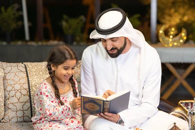 قراءة وتلاوة وحفظ القرآن الكريم، رجل عربي خليجي اماراتي يرتدي الكندورة والغترة  يمسك المصحف الشريف يجلس مع طفلته في الهواء الطلق في الخارج، تلاوة وقراءة كتاب الله تعالى ,الإجتهاد و الخشوع في العباده, التقرب الى رب العالمين في شهر رمضان المبارك، زينة واضاءة رمضانية