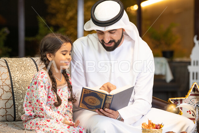 ترتيل وتلاوة وحفظ القرآن الكريم، رجل عربي خليجي اماراتي يرتدي الكندورة والغترة  يمسك المصحف الشريف يجلس مع طفلته في الهواء الطلق في الخارج، تلاوة وقراءة كتاب الله تعالى ,الإجتهاد و الخشوع في العباده, التقرب الى رب العالمين في شهر رمضان المبارك، زينة واضاءة رمضانية