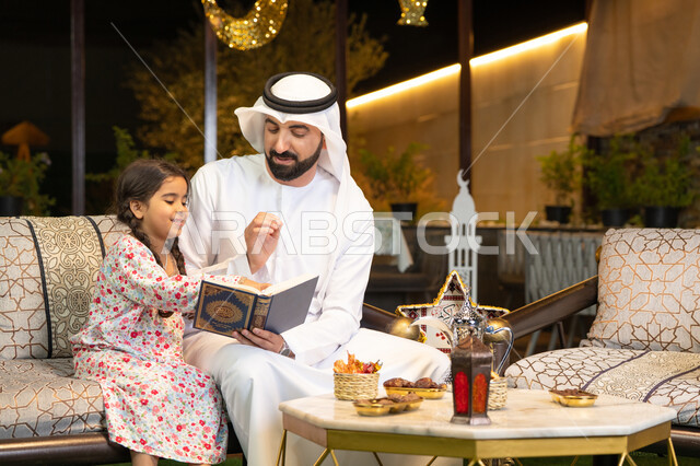 Reading, reciting and memorizing the Holy Quran, an Emirati Gulf Arab man wearing a kandura and ghutra holding the Holy Quran sitting with his daughter in the open air outside, reciting and reading the Book of God Almighty, diligence and humility in worship, getting closer to the Lord of the Worlds in the blessed month of Ramadan, Ramadan decorations and lighting