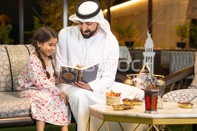 Chanting, reciting and memorizing the Holy Quran, an Emirati Gulf Arab man wearing a kandura and ghutra holding the Holy Quran sitting with his daughter in the open air outside, reciting and reading the Book of God Almighty, diligence and humility in worship, getting closer to the Lord of the Worlds in the blessed month of Ramadan, Ramadan decorations and lighting