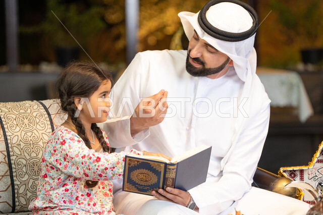 Reading, reciting and memorizing the Holy Quran, an Emirati Gulf Arab man wearing a kandura and ghutra holding the Holy Quran sitting with his daughter in the open air outside, reciting and reading the Book of God Almighty, diligence and humility in worship, getting closer to the Lord of the Worlds in the blessed month of Ramadan, Ramadan decorations and ighting