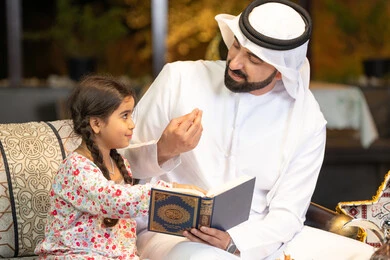 شرح وتلاوة وحفظ القرآن الكريم، رجل عربي خليجي اماراتي يرتدي الكندورة والغترة  يمسك المصحف الشريف يجلس مع طفلته في الهواء الطلق في الخارج، تلاوة وقراءة كتاب الله تعالى ,الإجتهاد و الخشوع في العباده, التقرب الى رب العالمين في شهر رمضان المبارك، زينة واضاءة رمضانية