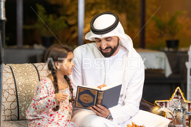 Chanting, reciting and memorizing the Holy Quran, an Emirati Gulf Arab man wearing a kandura and ghutra holding the Holy Quran sitting with his daughter in the open air outside, reciting and reading the Book of God Almighty, diligence and humility in worship, getting closer to the Lord of the Worlds in the blessed month of Ramadan, Ramadan decorations and lighting