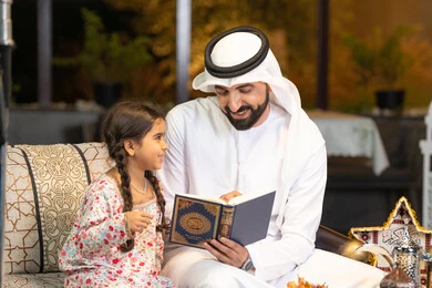 Chanting, reciting and memorizing the Holy Quran, an Emirati Gulf Arab man wearing a kandura and ghutra holding the Holy Quran sitting with his daughter in the open air outside, reciting and reading the Book of God Almighty, diligence and humility in worship, getting closer to the Lord of the Worlds in the blessed month of Ramadan, Ramadan decorations and lighting
