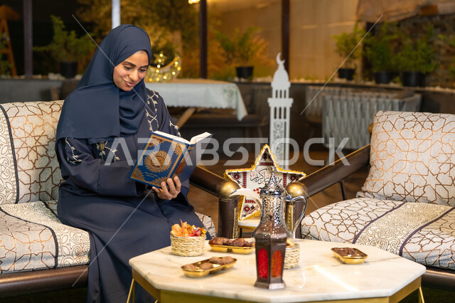Reciting and reading the Book of God Almighty, diligence and humility in worship, a Saudi Arabian Gulf woman wearing the hijab and abaya holding the Holy Quran in her hand, an Emirati woman sitting on a couch in the open air, getting closer to the Lord of the Worlds in the holy month of Ramadan, Ramadan decorations and lighting