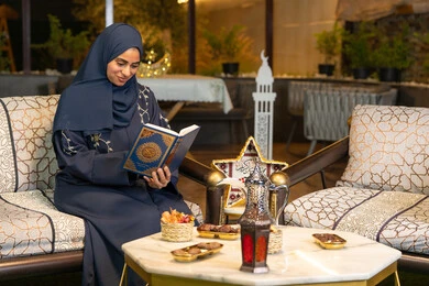 Reciting and reading the Book of God Almighty, diligence and humility in worship, a Saudi Arabian Gulf woman wearing the hijab and abaya holding the Holy Quran in her hand, an Emirati woman sitting on a couch in the open air, getting closer to the Lord of the Worlds in the holy month of Ramadan, Ramadan decorations and lighting