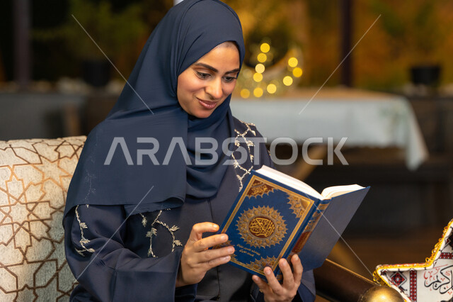 قراءة وتلاوة كتاب الله تعالى ,الإجتهاد و الخشوع في العباده, امرأة عربية سعودية خليجية ترتدي الحجاب و العباءة تمسك بيدها القرآن الكريم، سيدة اماراتية تجلس على الكنبة في الهواء الطلق, التقرب الى رب العالمين في شهر رمضان المبارك، زينة واضاءة رمضانية