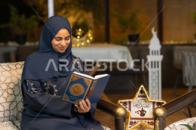 Reciting and reading the Book of God Almighty, diligence and humility in worship, a Saudi Arabian Gulf woman wearing the hijab and abaya holding the Holy Quran in her hand, an Emirati woman sitting on a couch in the open air, getting closer to the Lord of the Worlds in the holy month of Ramadan, Ramadan decorations and lighting