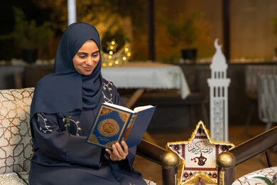 تلاوة وقراءة كتاب الله تعالى ,الإجتهاد و الخشوع في العباده, امرأة عربية سعودية خليجية ترتدي الحجاب و العباءة تمسك بيدها القرآن الكريم، سيدة اماراتية تجلس على الكنبة في الهواء الطلق, التقرب الى رب العالمين في شهر رمضان المبارك، زينة واضاءة رمضانية