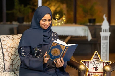Reciting and reading the Book of God Almighty, diligence and humility in worship, a Saudi Arabian Gulf woman wearing the hijab and abaya holding the Holy Quran in her hand, an Emirati woman sitting on a couch in the open air, getting closer to the Lord of the Worlds in the holy month of Ramadan, Ramadan decorations and lighting