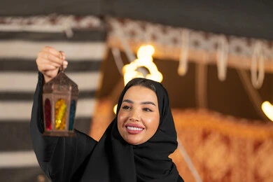 امرأه عربيه خليجيه سعوديه مبتسمه تحتفل بقدوم شهر رمضان المبارك, صوره مقربه لسيده اماراتيه ترتدي العباءه والحجاب الاسود تمسك بفانوس معدني ملون, خيمه مليئه بزينه وديكورات رمضانيه, التخييم الليلي الهادئ