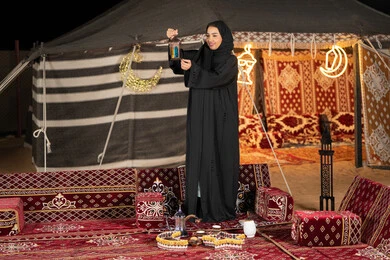 امرأه عربيه خليجيه سعوديه مبتسمه تحتفل بقدوم شهر رمضان المبارك, سيده اماراتيه ترتدي العباءه والحجاب الاسود تمسك بفانوس معدني ملون, خيمه مليئه بزينه وديكورات رمضانيه, التخييم الليلي الهادئ