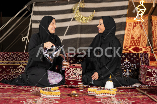 العادات والتقاليد الأصيلة في تقديم المشروبات الساخنة، حسن الضيافة والاستقبال عند العرب، مفهوم الأنوثة والنعومة، امرأة عربية خليجية سعودية ترتدي العباءة السوداء تقوم بصب القهوة في فنجان لصديقتها، سيدتان اماراتيتان تستمتعان بتناول القهوه