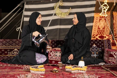 العادات والتقاليد الأصيلة في تقديم المشروبات الساخنة، حسن الضيافة والاستقبال عند العرب، مفهوم الأنوثة والنعومة، امرأة عربية خليجية سعودية ترتدي العباءة السوداء تقوم بصب القهوة في فنجان لصديقتها، سيدتان اماراتيتان تستمتعان بتناول القهوه