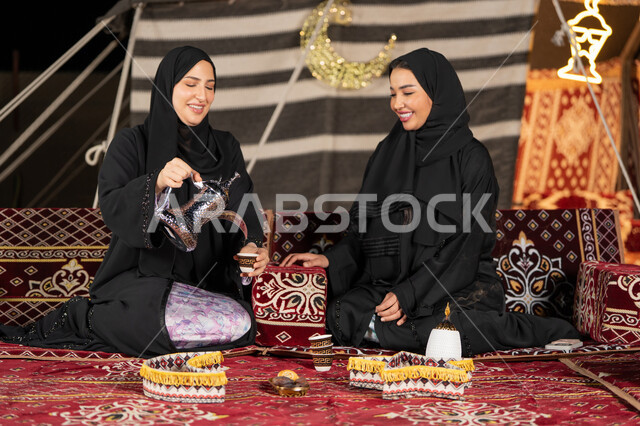 العادات والتقاليد الأصيلة في تقديم المشروبات الساخنة، حسن الضيافة والاستقبال عند العرب، مفهوم الأنوثة والنعومة، امرأة عربية خليجية سعودية ترتدي العباءة السوداء تقوم بصب القهوة في فنجان لصديقتها، سيدتان اماراتيتان تستمتعان بتناول القهوه