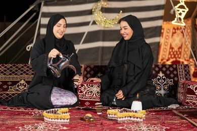 العادات والتقاليد الأصيلة في تقديم المشروبات الساخنة، حسن الضيافة والاستقبال عند العرب، مفهوم الأنوثة والنعومة، امرأة عربية خليجية سعودية ترتدي العباءة السوداء تقوم بصب القهوة في فنجان لصديقتها، سيدتان اماراتيتان تستمتعان بتناول القهوه