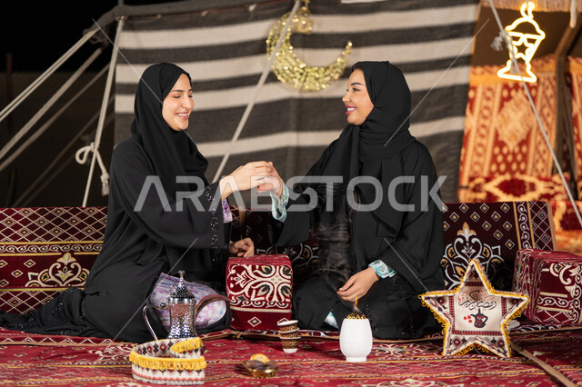 حسن الضيافة والاستقبال عند العرب، العادات والتقاليد الأصيلة في تقديم المشروبات الساخنة،  مفهوم الأنوثة والنعومة، امرأة عربية خليجية سعودية ترتدي العباءة السوداء تقوم بصب القهوة في فنجان لصديقتها، سيدتان اماراتيتان تستمتعان بتناول القهوه