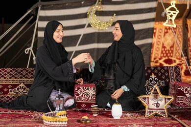 حسن الضيافة والاستقبال عند العرب، العادات والتقاليد الأصيلة في تقديم المشروبات الساخنة،  مفهوم الأنوثة والنعومة، امرأة عربية خليجية سعودية ترتدي العباءة السوداء تقوم بصب القهوة في فنجان لصديقتها، سيدتان اماراتيتان تستمتعان بتناول القهوه