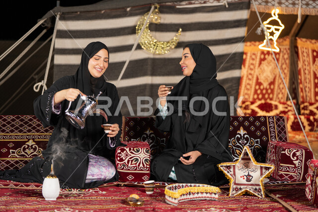 العادات والتقاليد الأصيلة في تقديم المشروبات الساخنة، حسن الضيافة والاستقبال عند العرب، مفهوم الأنوثة والنعومة، امرأة عربية خليجية سعودية ترتدي العباءة السوداء تقوم بصب القهوة في فنجان لصديقتها، سيدتان اماراتيتان تستمتعان بتناول القهوه