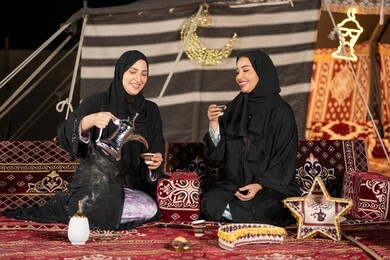 العادات والتقاليد الأصيلة في تقديم المشروبات الساخنة، حسن الضيافة والاستقبال عند العرب، مفهوم الأنوثة والنعومة، امرأة عربية خليجية سعودية ترتدي العباءة السوداء تقوم بصب القهوة في فنجان لصديقتها، سيدتان اماراتيتان تستمتعان بتناول القهوه