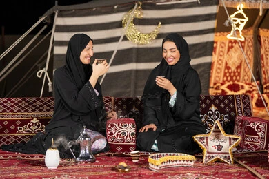 حسن الضيافة والاستقبال عند العرب، العادات والتقاليد الأصيلة في تقديم المشروبات الساخنة،  مفهوم الأنوثة والنعومة، امرأة عربية خليجية سعودية ترتدي العباءة السوداء تقوم بصب القهوة في فنجان لصديقتها، سيدتان اماراتيتان تستمتعان بتناول القهوه