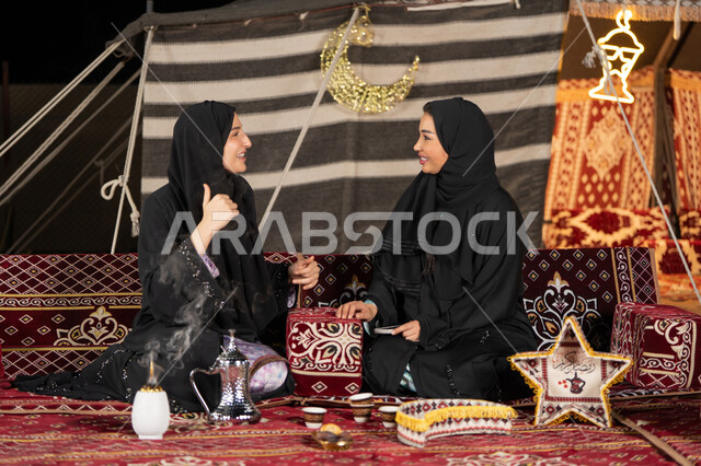 Talking and chatting about happy topics, two Arab Gulf women talking about a topic, using the latest modern technologies, two Saudi women wearing the abaya and hijab sitting in the tent at night, the origins of the popular council