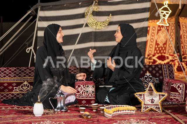 Talking and chatting about happy topics, two Arab Gulf women talking about a topic, using the latest modern technologies, two Saudi women wearing the abaya and hijab sitting in the tent at night, the origins of the popular council